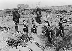 Five soldiers dig a trench, supervised by two standing above them.