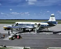 A white and blue aeroplane on a runway, surrounded by tankers of fuel and other equipment. The tail bears a stylised red Zimbabwe Bird, and the words "AIR RHODESIA" are painted above the windows of the cabin. Towards the rear are painted the letters "VP-YNC", with the green and white Rhodesian flag rendered above.