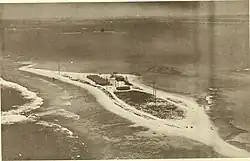 Aerial black-and-white photograph of a small, narrow atoll island surrounded by shallow ocean waters. The island has a few low buildings and utility poles, with sparse vegetation and sandy shores. Gentle waves break along the left side, and some rocky outcroppings are visible in the surrounding waters.
