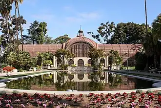 Photograph of the Botanic Building, one of several buildings in Balboa Park. A red conservatory building, it is fronted by a reflecting pool and surrounded by multiple species of trees.
