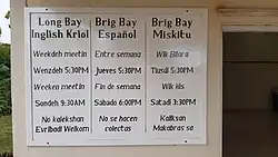 Photograph of a board on a wall with three columns. The column on the left says "Long Bay / Inglish Kriol. Weekdeh meetin / Wenzdeh 5:30PM / Weeken meetin / Sondeh 9:30AM / No kalekshan / Evribadi Welkom". The column in the middle is in Spanish. The column on the right is in Miskito. To the right, an open door. To the left, a bit of greenery.
