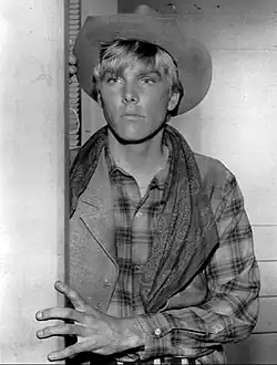A black-and-white photo of a young white man with light-colored hair. He is wearing a cowboy hat and flannel shirt. He looks to the left of the camera, holding his left hand against the wall.