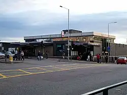 A building with a blue sign reading "BLACKHORSE ROAD STATION" in white letters and several people and cars in the foreground all under a blue sky