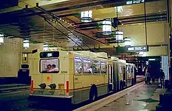 A white bus with raised poles stopped at the curb of an underground station. The granite-tiled floors reflect bright lanterns placed above the platform, adjacent to an upper level walkway.