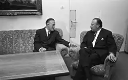Two middle-aged men sitting in a sofa and a chair around the corner of an empty table