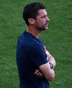 A middle-aged man wearing a blue t-shirt stands with his arms crossed.