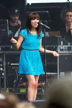 A Caucasian female with shoulder-length, wavy brown hair and wearing a powder blue dress sings and pearls into a microphone on stage, extending both arms in an emphatic gesture.
