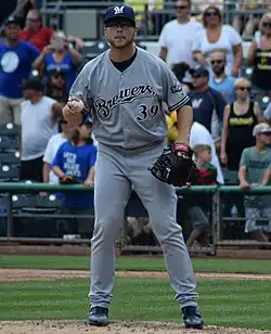 A man in a gray baseball uniform with "Brewers" across the chest and a navy cap standing on a pitcher's mound with a ball in one hand