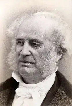 Bust portrait photograph of an old man, mostly balded with whispy white hair at the edges of his head and mutton chops, wearing a high-collared dress shirt.