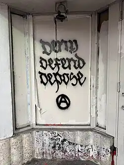 Stencil graffiti reading "Deny Defend Depose" with gothic black letters and the Circle-A anachist symbol below on a covered window in Miami Beach, Florida.
