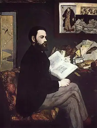 A man sits engrossed in a book, wearing a dark suit and light trousers, with a beard and dark hair. Behind him, framed artworks adorn the wall, including Manet's Olympia and a Japanese print.