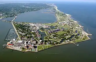 An aerial view of a flat promontory of land with a large fortification in the center. There are many modern buildings and roads surrounding the fort. On either side of the promontory are wide expanses of water.