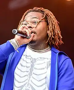 A man with brownish dreadlocks in a blue zip through sweat top and white shirt holding a microphone