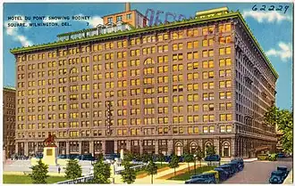 Postcard of a black and white photo with hand-coloring showing the building in front of a square with antique cars parked throughout