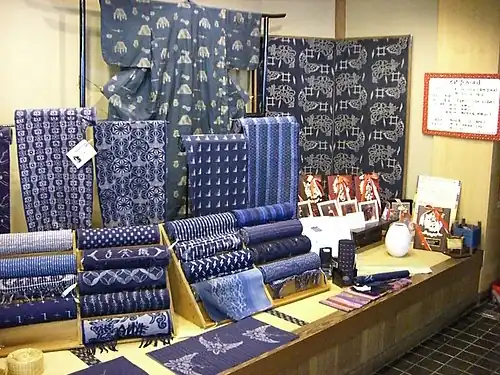 A display table showing a number of different indigo-dyed kasuri fabrics, some stacked on wooden stands in roll-form, others displayed flat in front of the rolls, and some hanging on small stands.