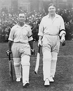 Two batsmen walk on to a cricket ground with a crowd behind them