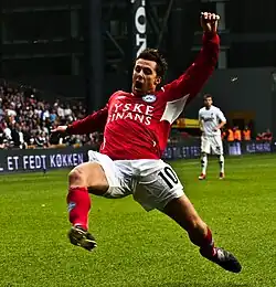 A photograph of a man dressed in a red football sweater, white sport shorts and red socks. The man is in mid play during a football match throwing himself forward for something or someone who is off-picture.