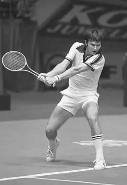 A brown-haired man dressed in a white shirt swings a two-handed backhand