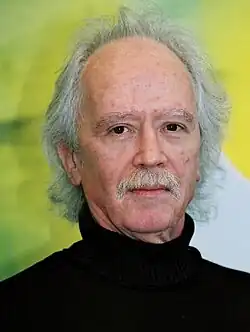 An elderly white man with a gray mustache and gray receding hair faces the camera with a neutral expression.