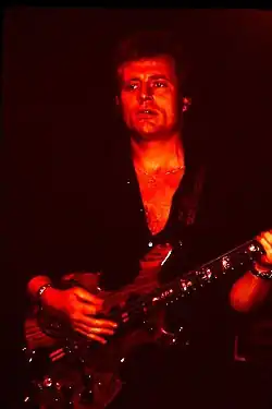 A red-tinged photograph of John Paul Jones playing an 8-string bass guitar in concert