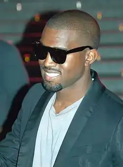 An Afro-American man with a ponytail on top of her head is wearing a black leather jacket before a light-coloured background.