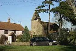 Stone church with square tower in the background partially obscured by trees. To the left is a pink painted house with red roof and in the foreground a car and grass area.