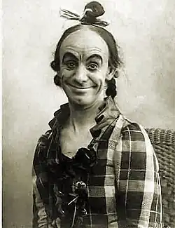 Head and torso photo of a man in women's clothing, sitting. He is smiling at the viewer with mouth closed but a wide smile, and eyebrows raised high, as is typical in the photos of Leno in character
