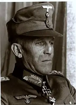 A grim looking man wearing a peaked cap and a military uniform with an Iron Cross displayed at his neck.