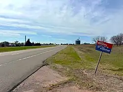 Welcome sign on highway into Manitou, 3-2025; grain elevator in background