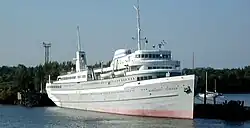 Starboard side of a gray-white boat with two lifeboats, four decks, and a small smokestack docked on a pier