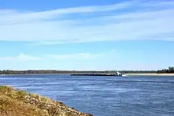 An open stretch of river water. A barge is in the center of the image, and in the background is a wooded riverbank. There are no signs of human habitation on the riverbanks.