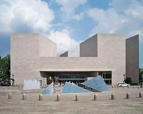 The exterior of the National Gallery East Building, a large gray building, rises above a stone plaza. Short square towers appear on either side of the building, and an array of irregular glass pyramids are in the middle of the plaza.