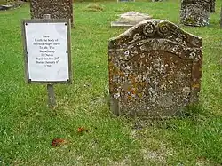 A small stone headstone, dotted with lichen, in a graveyard. The headstone has a simple carved frame, with a scroll design at the top, and an inscription in the Roman alphabet. Next to the headstone is a sign with a transcription of the inscription, which reads "Here lyeth the body of Myrtilla, negro slave to Mr. Thos Beauchamp of Nevis. Bapt. Oct. ye 20th. Buried Jan ye 6th, 1705"