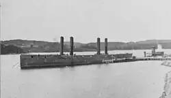 USS Nevada, a long-looking frigate, resting near a pier. She lacks sails and details above her main deck aside from 4 prominent funnels.