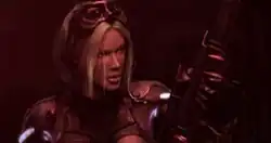 A young blonde woman in an armored suit loads a sniper rifle in a red lit room.