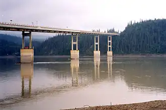 A beige bridge on supports spanning a brown colored river and evergreen hills in the background.