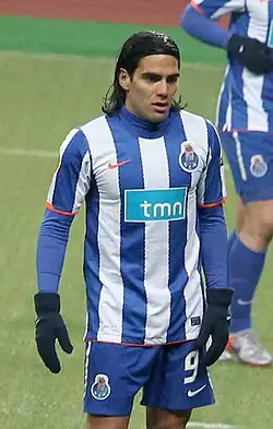 Man with long dark hair held with a headband, wearing a football kit composed of a shirt with vertical blue-and-white stripes and blue shorts. He also wears a blue sleeved undershirt and winter gloves.