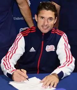 A man wearing a blue New England Revolution tracksuit is sitting and signing a piece of paper.