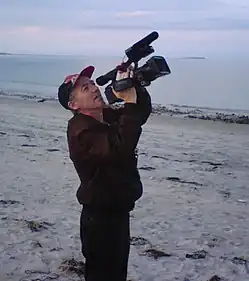 Photograph of Richard Dindo with camera, on a beach at Plymouth, Massachusetts, USA, while directing the film Mars Dreamers