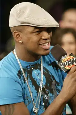An African-American man is wearing a blue T-shirt and white cap. He is talking on a microphone and smiling.