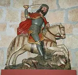 Photograph of a statue of a white bearded rider in armor wearing a pilgrim hat and a a red cape. He is raising his sword over his head. His white horse gallops over the fallen bodies of downscaled warrior with scimitars.