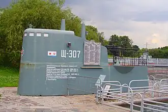 A color photograph of a blue/grey metallic structure (a submarine's tower) with Russian text and the Soviet flag on it, next to an enclosed pool