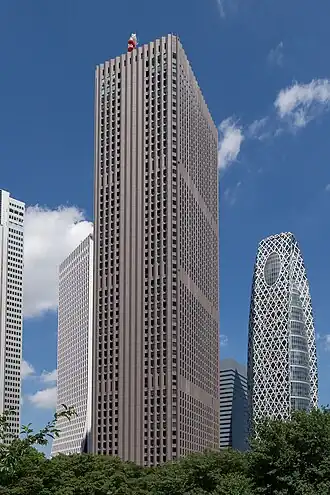 Ground-level view of a brown, rectangular high-rise; the window placement creates several horizontal bands on one side and one vertical stripe on the other