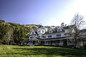 A white mansion house topped by a black roof stands amid green hills and forests.