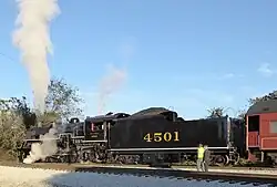 A black steam locomotive with tender coupled to a passenger coach, waiting to depart an out-of-view station