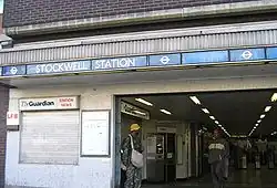 A brown-bricked building with a rectangular, light blue sign reading "STOCKWELL STATION" in white letters all under a light blue sky