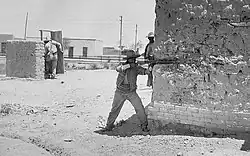 A man aims a rifle from around the corner of a bullet-scarred wall. In the background another man is attempting to take cover behind a low wall and partial door.