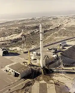 Photograph of the Thor-Agena vehicle on the launch pad at Space Launch Complex 10, surrounded by scrub vegetation and various support buildings, the Pacific Ocean in the distant background.