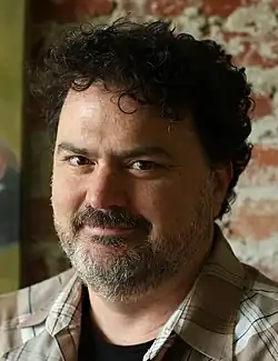 A portrait of a man with curly black hair and a salt-and-pepper beard facing the left of the screen, eyes turned towards the viewer