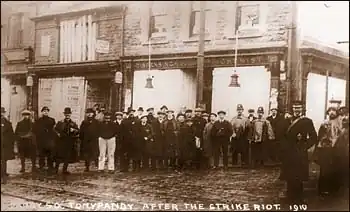A sepia-toned photograph shows a group of men standing in front of a row of buildings with signs indicating shops or businesses. The men are dressed in early 20th-century attire, including hats and overcoats. The image is labeled "Tonypandy after the strike riot 1910”. The men appear somber, and the buildings show signs of disrepair or damage.